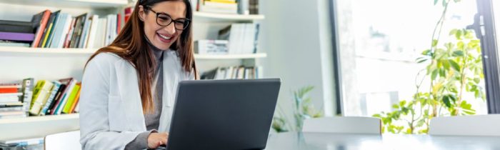 Female medical professional in a white coat smiling at her laptop in a modern office, surrounded by bookshelves and plants—engaged in cosmetic medicine online training or teleconsultation